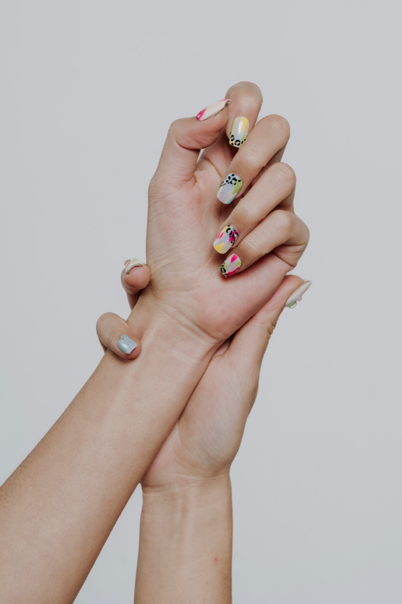 Two clasped hands with full sets of vibrant SNS nail art in pinks, yellows, greens and black leopard accents